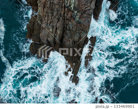 Waves crash against rugged cliffs under the bright sunlight of a clear day by the ocean Waves crash against rugged cliffs under the bright sunlight of a clear day by the ocean 133469901