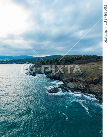 Waves crashing against rocky shoreline under a cloudy sky at dusk by the coast Waves crashing against rocky shoreline under a cloudy sky at dusk by the coast 133469903