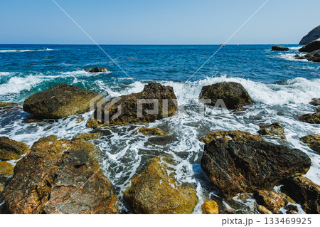 Tranquil waves crash against rugged rocks along a sunny coastal shore, capturing the beauty of nature's rhythm and serenity Tranquil waves crash against rugged rocks along a sunny coastal shore, capturing the beauty of nature's rhythm and serenity 133469925
