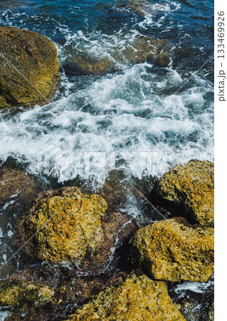 Waves crashing against rugged rocks on a sunny day by the seashore in a tranquil coastal setting Waves crashing against rugged rocks on a sunny day by the seashore in a tranquil coastal setting 133469926