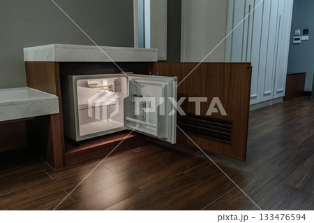 A small refrigerator sits inside a wooden cabinet with its door open, and soft interior light highlights the empty shelves and the bottle inside. 133476594