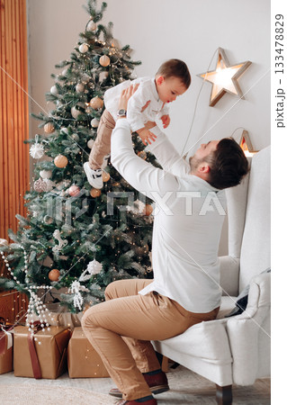 Father joyfully lifts his young son in front of a decorated Christmas tree with presents nearby during a festive holiday celebration at home 133478829