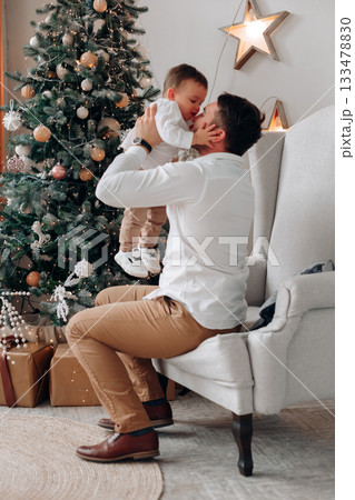 Father shares a joyful moment with his baby son in front of a decorated Christmas tree during a festive holiday celebration at home Father shares a joyful moment with his baby son in front of a decorated Christmas tree during a festive holiday celebration at home 133478830