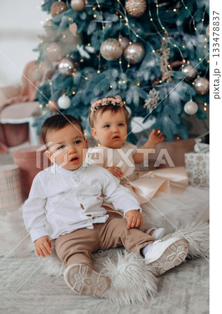 Children sitting on a soft rug next to a decorated Christmas tree in a cozy indoor space filled with holiday decorations and gifts 133478837