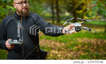 A bearded man in a black jacket stands outdoors, showcasing a small drone while holding a remote control. The surrounding park features vibrant autumn leaves. A bearded man in a black jacket stands outdoors, showcasing a small drone while holding a remote control. The surrounding park features vibrant autumn leaves. 133479786