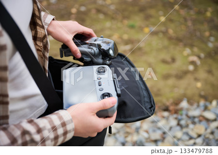 Holding a remote control, a person stands on the ground ready to pilot a drone above beautiful autumn scenery. Leaves scatter around as nature thrives. 133479788