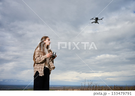 A young woman operates a drone in an expansive outdoor area with a cloudy sky. The scene captures a sense of adventure and technology in nature. 133479791