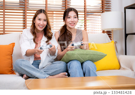 Two asian woman playing video games together with controllers in a cozy living room at home. 133481428
