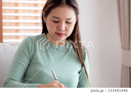 Happy young asian woman writing on book sitting on sofa with relax in living room at home. 133481446