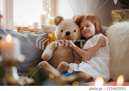 Small girl sitting on sofa indoors at home at Christmas, hugging teddy bear. Small girl sitting on sofa indoors at home at Christmas, hugging teddy bear. 133482033