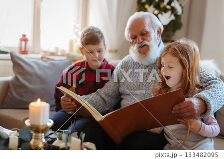 Small children with senior grandfather indoors at home at Christmas, looking at photographs. 133482095