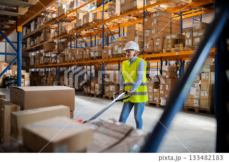 Older woman as warehouse worker pulling a pallet truck with boxes. 133482183