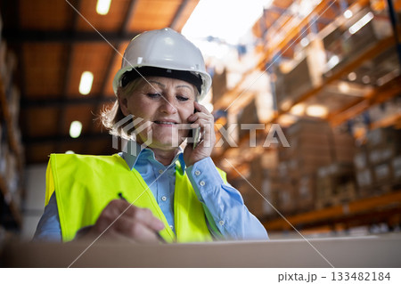 Female warehouse manager or supervisor with smartphone, making phone call. Female warehouse manager or supervisor with smartphone, making phone call. 133482184