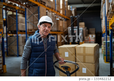 Older male warehouse worker pulling a pallet truck. Older male warehouse worker pulling a pallet truck. 133482212