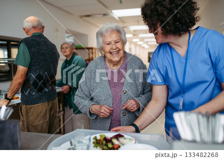 Senior woman having lunch with supportive caregiver in community center cafeteria. Senior woman having lunch with supportive caregiver in community center cafeteria. 133482268