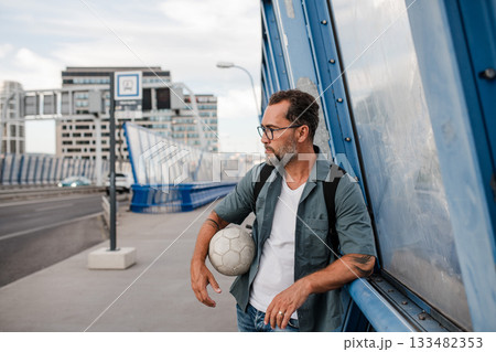 Bearded man with ball under arm using phone while waiting for bus. 133482353