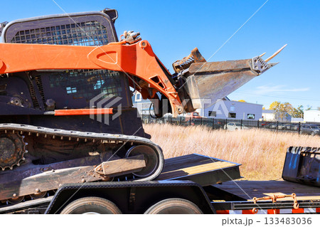 Skid steer machinery loader is positioned on flatbed trailer under its equipment. Skid steer machinery loader is positioned on flatbed trailer under its equipment. 133483036