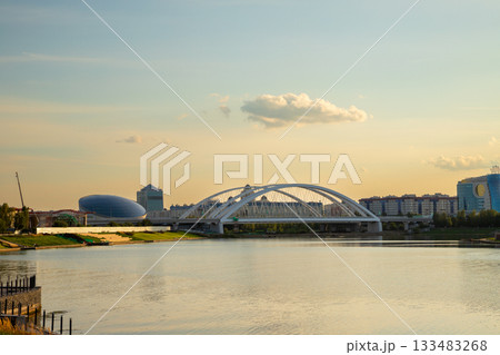 A vibrant sunset casts warm colors over the riverbank, showcasing a modern white bridge, buildings, and the calm water reflecting the evening sky. Astana, Kazakhstan, Argar Bridge over the Ishim River 133483268