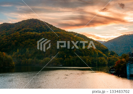 mountainous landscape with lake tarnita in autumn at dusk. forest on hills near the shore of somes river under orange sky. beautiful view in apuseni nature park romania, europe. stunning place mountainous landscape with lake tarnita in autumn at dusk. forest on hills near the shore of somes river under orange sky. beautiful view in apuseni nature park romania, europe. stunning place 133483702