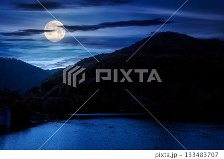mountainous landscape with lake in autumn at night. forest on hills near the shore of river under dark sky in full moon light. beautiful view of stunning place 133483707