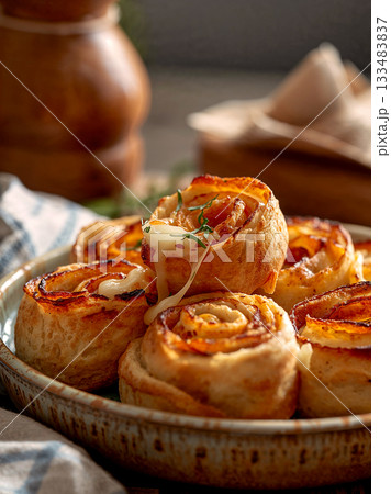 Close up of delicious savory puff pastry rolls with melted cheese and herbs. Golden brown baked pinwheel snacks on a rustic plate. 133483837