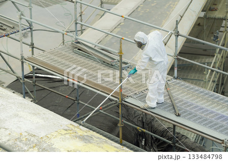 Worker in protective gear conducts cleaning task on scaffold above a waterway near construction site during daylight hours Worker in protective gear conducts cleaning task on scaffold above a waterway near construction site during daylight hours 133484798