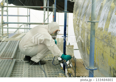 Worker in protective gear sprays paint on large metal tank while crouching on scaffolding at industrial site during daylight hours 133484801