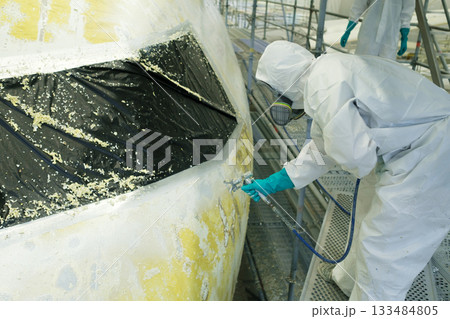 Professional worker applies spray paint to an aircraft in a hangar during repairs, ensuring precision and safety while wearing protective gear 133484805