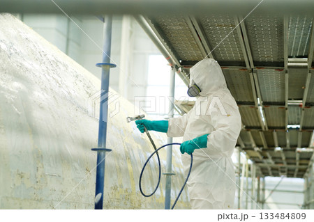 Worker in protective gear spray painting a large surface on scaffolding in an industrial setting during daylight hours Worker in protective gear spray painting a large surface on scaffolding in an industrial setting during daylight hours 133484809
