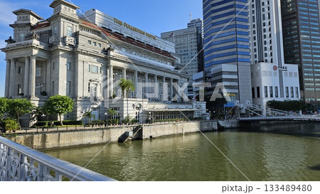 The grand Fullerton Hotel stands majestically by the riverside, framed by lush trees and modern skyscrapers under a bright blue sky, creating a striking blend of heritage architecture The grand Fullerton Hotel stands majestically by the riverside, framed by lush trees and modern skyscrapers under a bright blue sky, creating a striking blend of heritage architecture 133489480