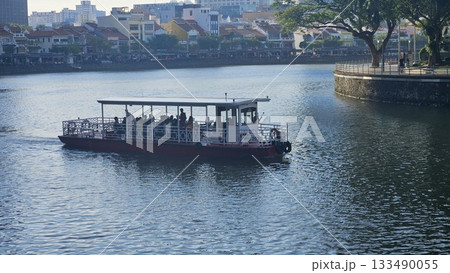 A riverboat filled with passengers glides calmly across the reflective water as colorful riverside buildings and lush trees create a peaceful and scenic urban atmosphere in the warm afternoon light. 133490055