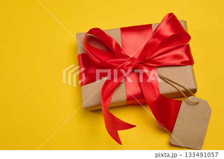 A gift box with a red silk bow and a empty brown tag on a yellow background, top view A gift box with a red silk bow and a empty brown tag on a yellow background, top view 133491057