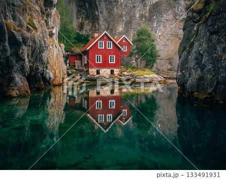 A stunning image of a vivid red wooden house and cottage nestled between steep rocky cliffs, perfectly reflected in the turquoise-green water of a calm fjord or lake. 133491931