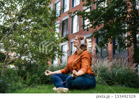 Beautiful mid size woman meditating in park, listening podcast. 133492329