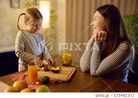 Orange juice on table while daughter cuts fruit and mother watches with warm smile 133492351