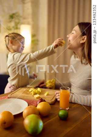 Orange juice on table while daughter feeds fruit to smiling mother in warm playful mood 133492352