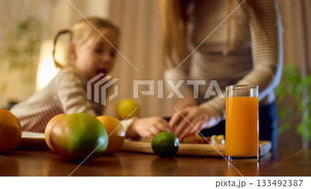 Bright orange juice standing on table during fruit preparation with joyful little girl and her mom Bright orange juice standing on table during fruit preparation with joyful little girl and her mom 133492387