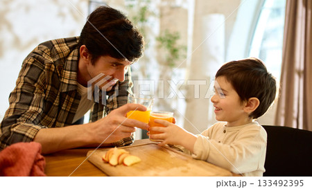 Orange juice glasses in father and son hands during warm cheerful kitchen toast moment 133492395