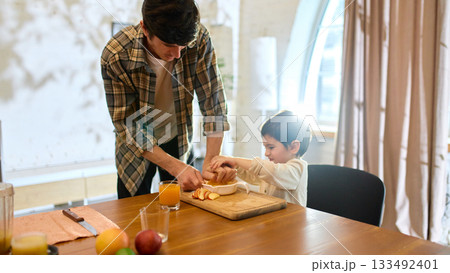 Orange juice in glass with father and son squeezing citrus together in bright kitchen light 133492401