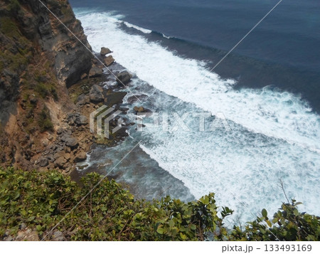 Ocean Waves Crashing Against a Cliffside with Rocky Shores and Greenery Under a Clear Sky 133493169