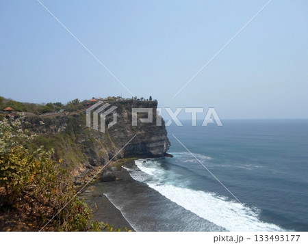 A Cliffside Shoreline with Waves Crashing Against Rocks and Greenery on the Cliff Under a Clear Sky A Cliffside Shoreline with Waves Crashing Against Rocks and Greenery on the Cliff Under a Clear Sky 133493177