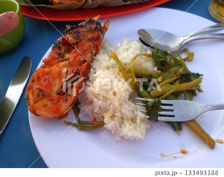 A Plate of Grilled Lobster with Rice and Sauteed Vegetables on a Beachside Table with Utensils 133493188