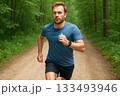 Man Running on Forest Trail Wearing Blue Shirt and Black Shorts Surrounded by Lush Green Trees 133493946