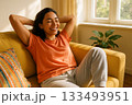 Woman Relaxing on Yellow Sofa with Arms Behind Head in Sunlit Living Room Featuring Plants and Window 133493951