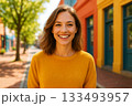 Woman Wearing Yellow Sweater Smiling Brightly on Colorful Street with Trees and Buildings in Background 133493957