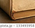 Close-Up of Old Book Corner with Worn Brown Cover and Torn Edge on Wooden Table Surface 133493958