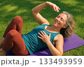 Woman Practicing Relaxation Exercise on Purple Yoga Mat in Sunlit Grass with Eyes Closed and Smile 133493959