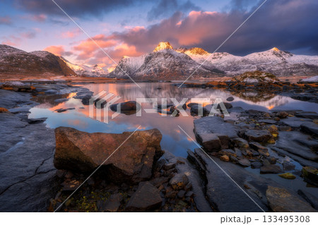 Snowy mountains reflecting on calm lake with stones at sunrise 133495308