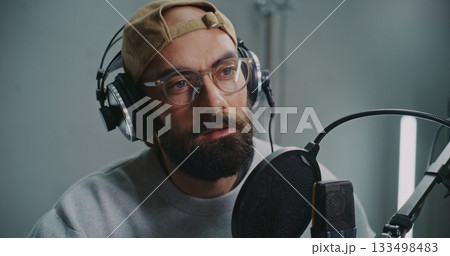Expressive Bearded Podcast Host Wearing Cap and Glasses Talks Into High Quality Microphone Expressive Bearded Podcast Host Wearing Cap and Glasses Talks Into High Quality Microphone 133498483