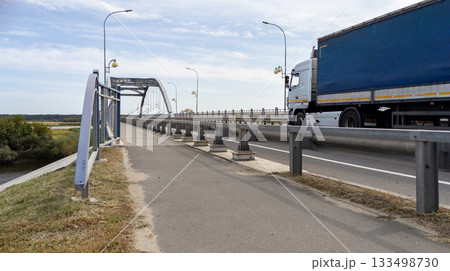 Large blue truck drives along a road across a river bridge against the backdrop of cloudy sky Large blue truck drives along a road across a river bridge against the backdrop of cloudy sky 133498730